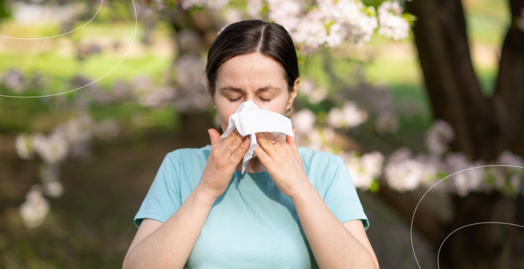 Mujer al aire libre sonándose la nariz con un pañuelo de papel frente a árboles en flor debido a síntomas de alergia estacional.